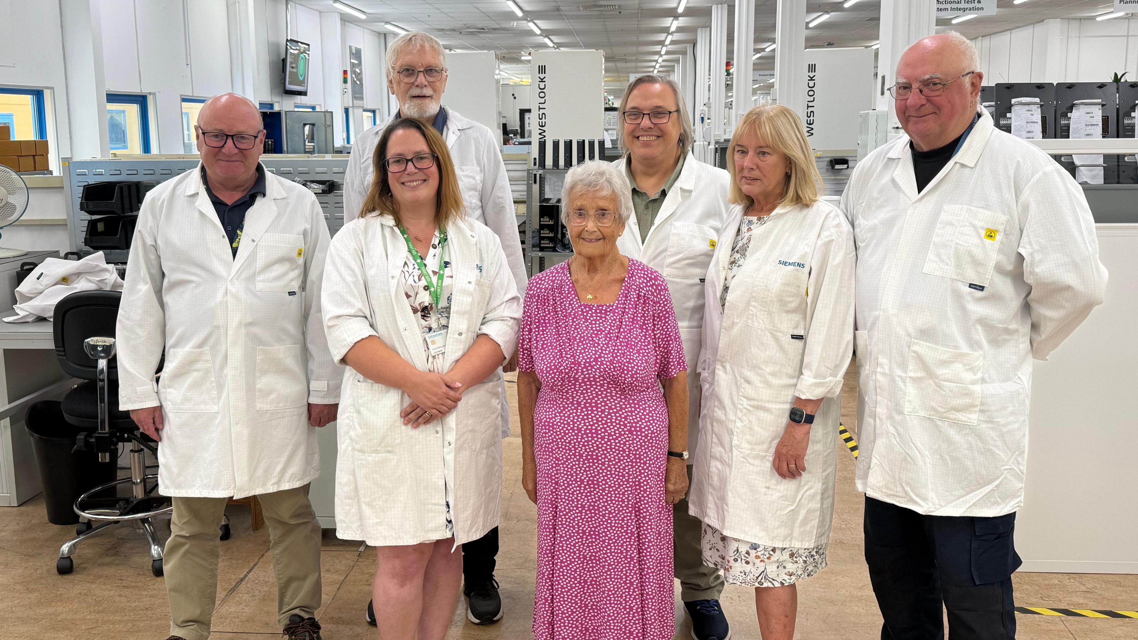 An elderly woman wearing a pink dress stands in the middle of a group of seven people. The others, all taller than her, are wearing lab coats. They are standing in an engineering space with workspaces, screens, chairs and a desktop fan all visible in the background.