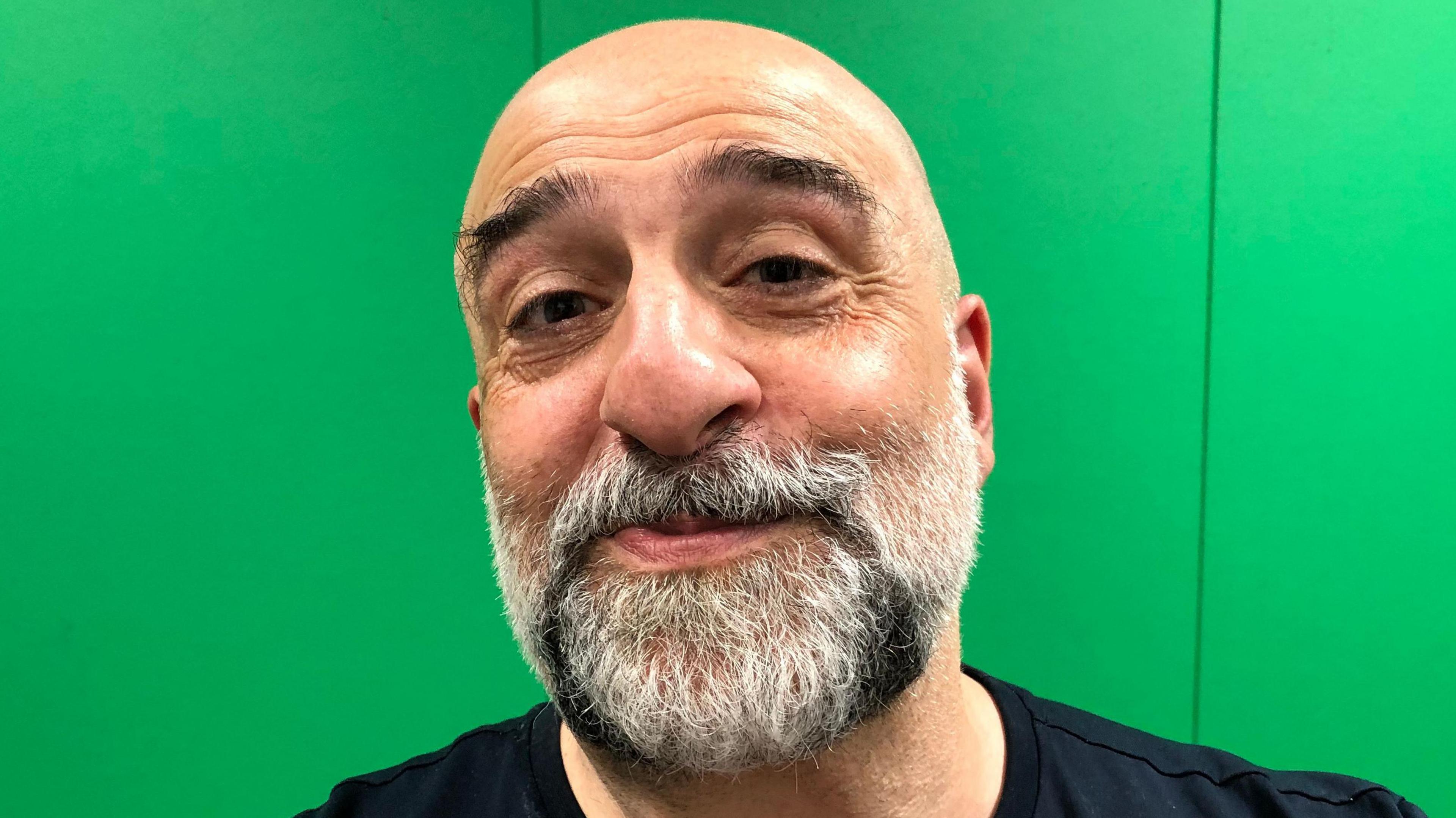 A photo of actor and comedian Omid Djalili in the BBC Suffolk studio. He is facing the camera and smiling in front of a green background. He is bald and has a black and white beard. He is wearing a black t-shirt.