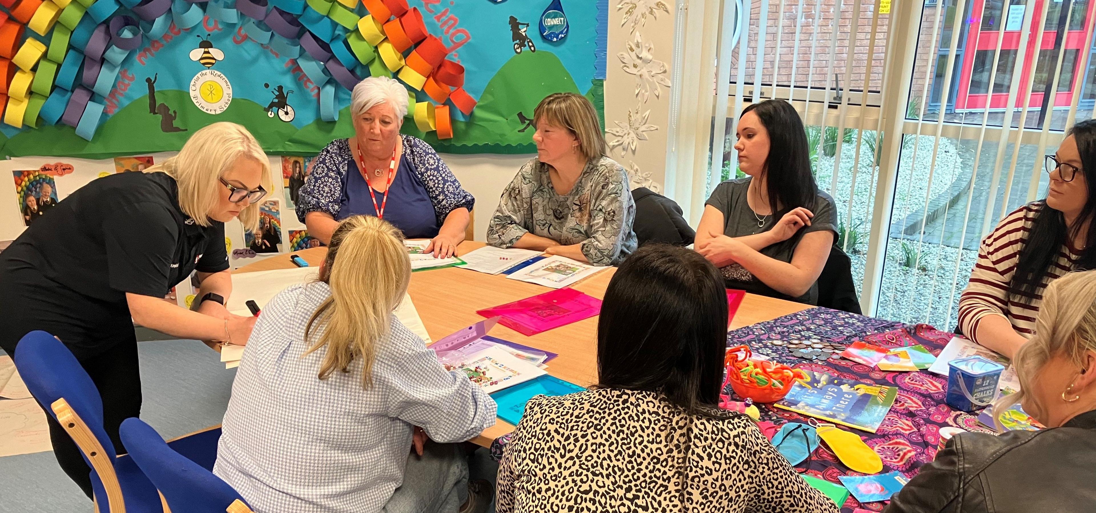 Five adults watch on as a teacher at Christ the Redeemer Primary School in Dunmurry writes with a marker on a sheet of paper. The teacher, leaning over a desk, is wearing glasses with a black t-shirt. There are colourful patterns and folders on the table as well as a textbook, markers and a small blue box.