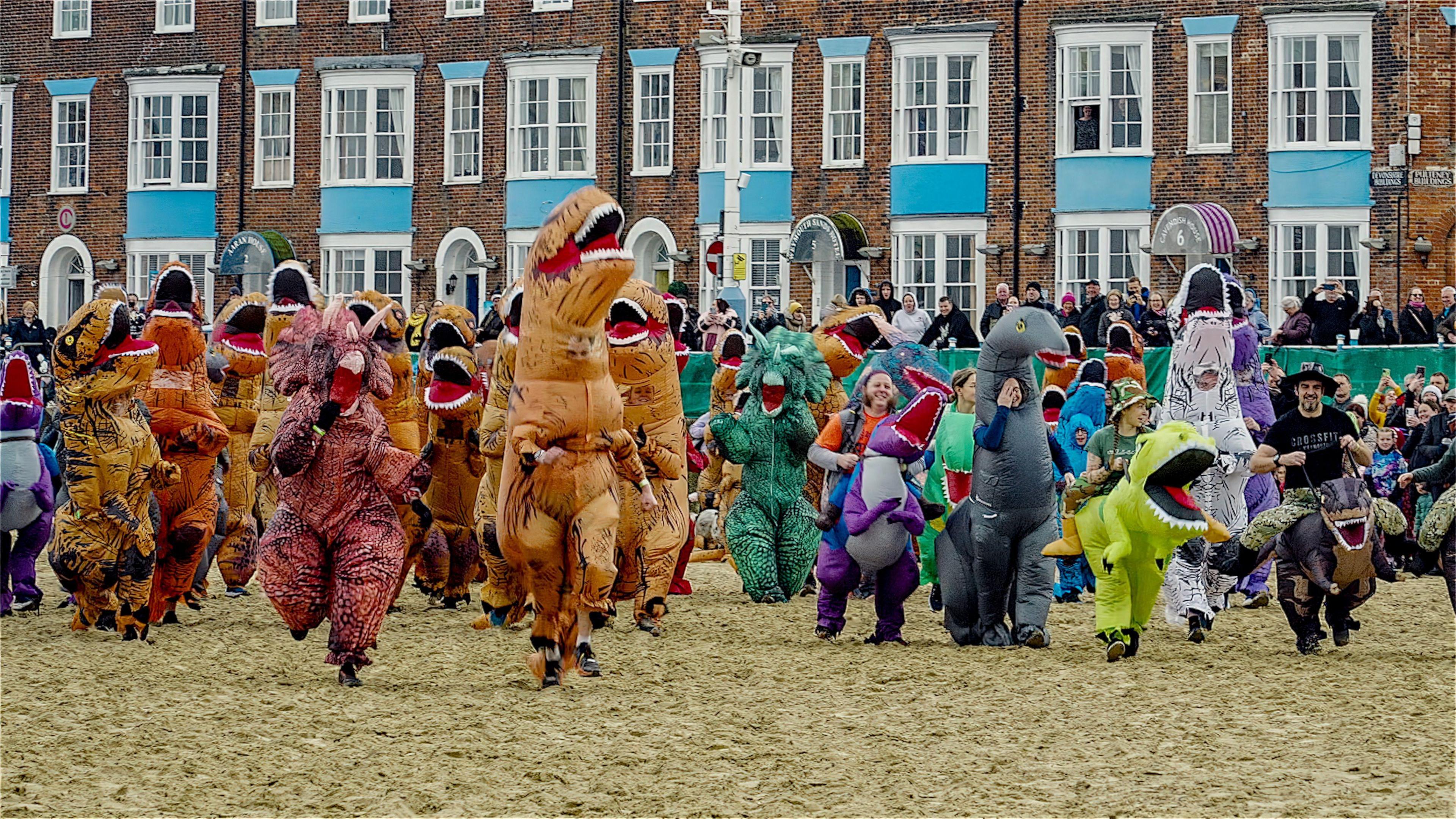 A group of people wearing dinosaur costumes running along a beach. Spectators are watching from a promenade behind them, with a row of brick buildings behind them.