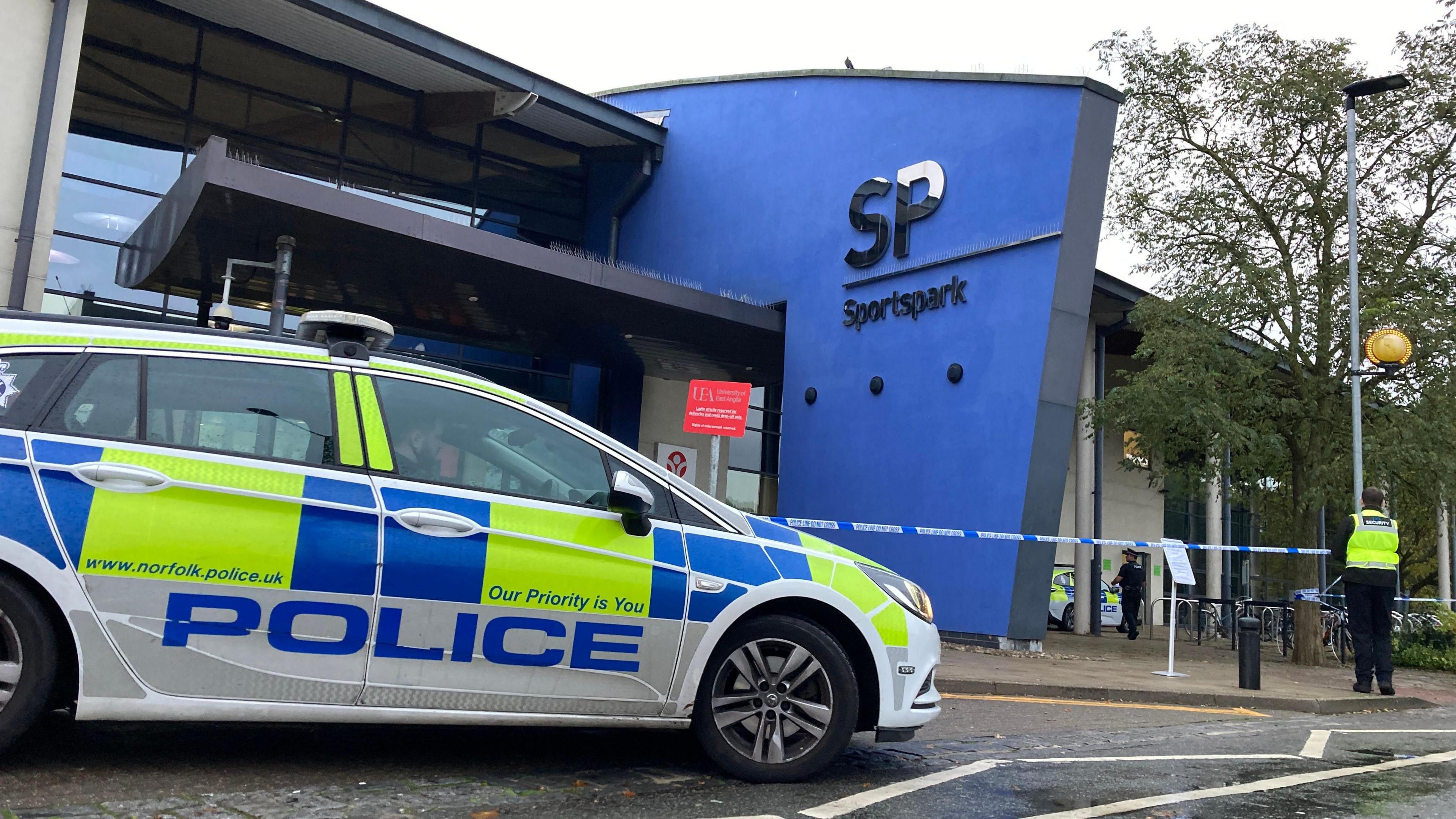 A police officers stands in front of police tape at the Sportspark, which is a large blue and white building. There is a police car at the front of the picture which is also in front of the building.