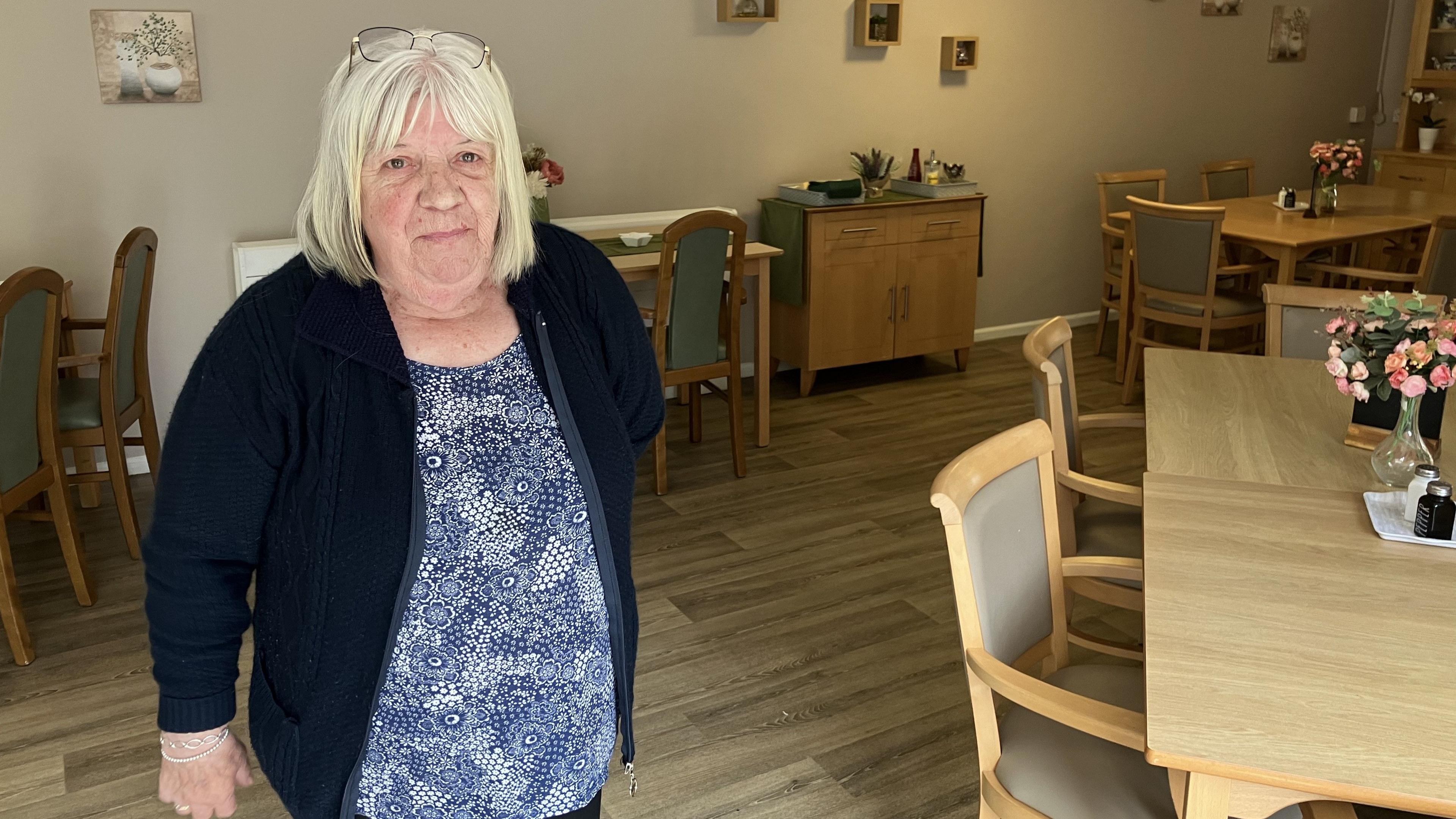 Pat stood in the dining room, surrounded by tables with flowers on