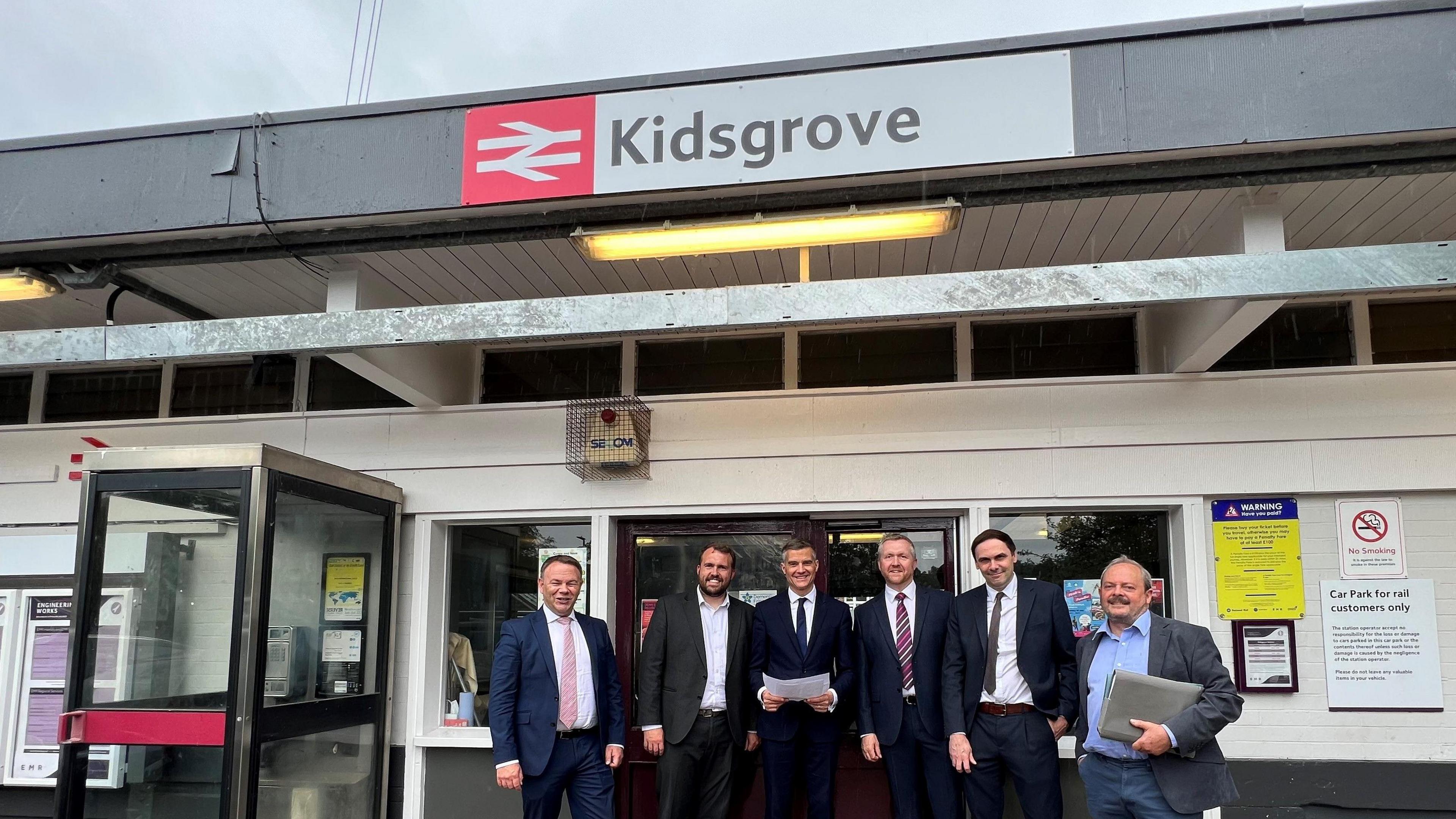 A group of six men wearing suits are stood on the station concourse of Kidsgrove Railway Station. There is a phone box to the left hand side of the image, and railway signage on the building behind them.