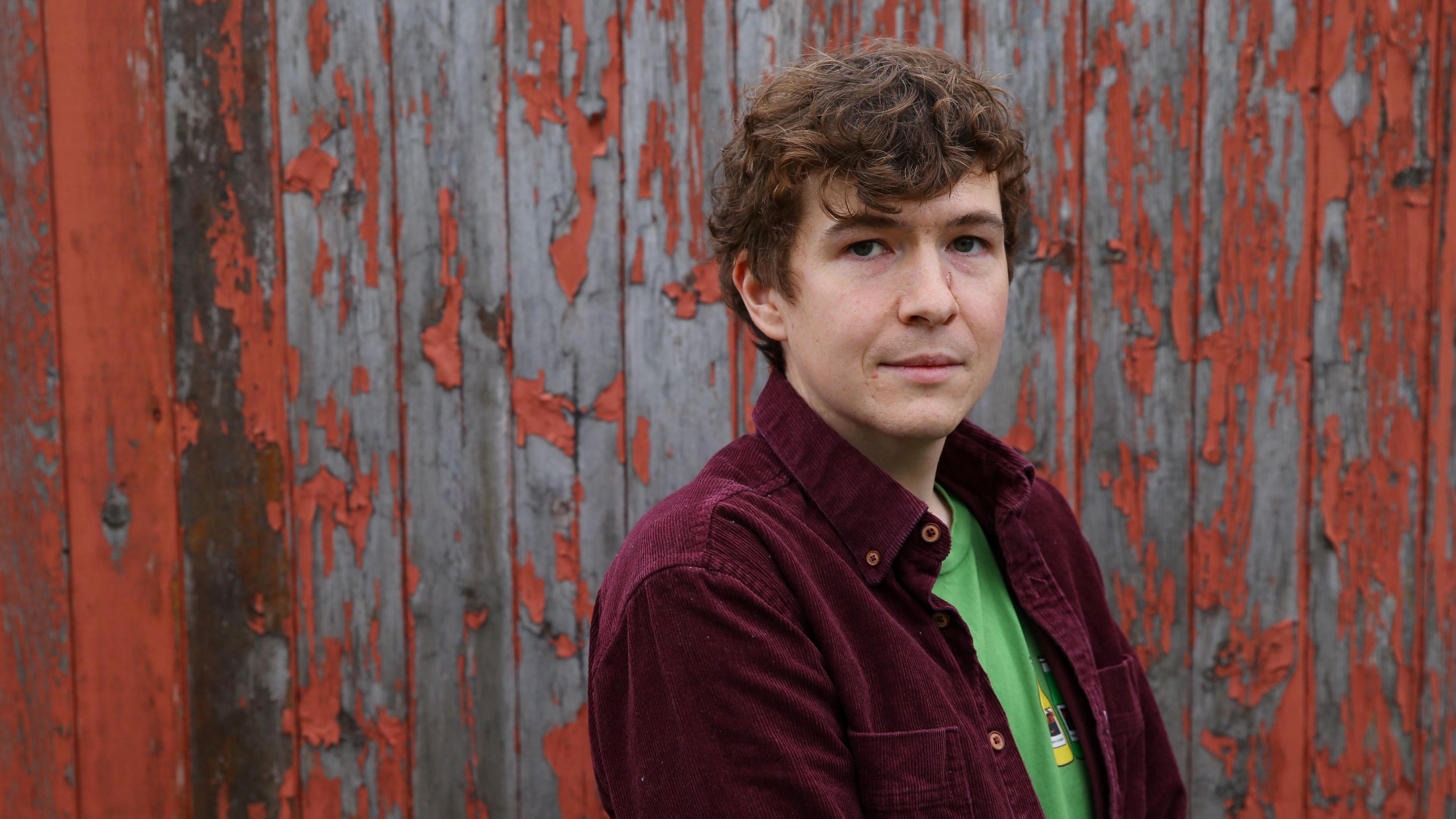Novelist Callum McSorley, a brown-haired man in a purple cord shirt and green T-shirt looks along his shoulder, standing side-on.
