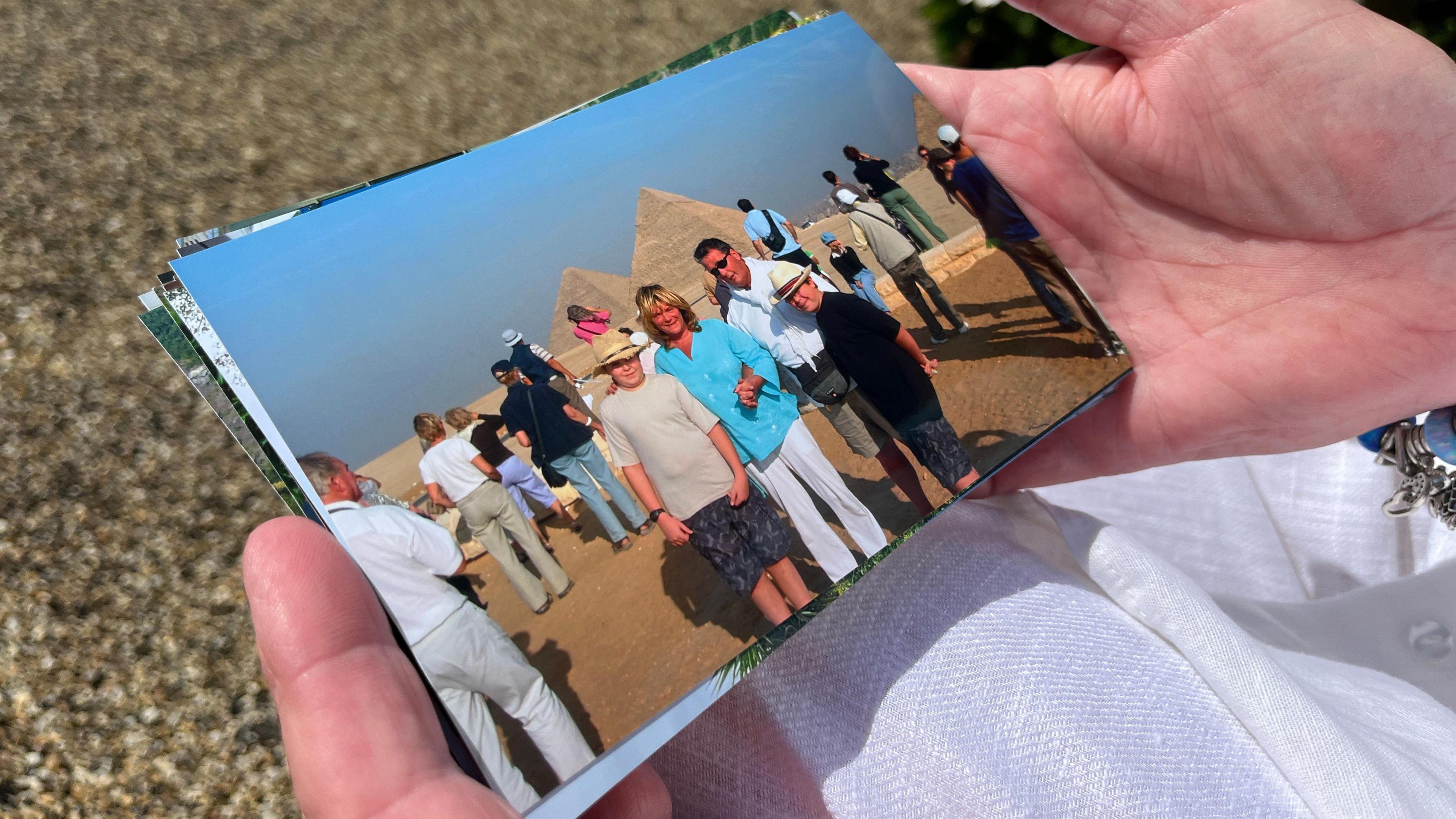 Alison holds a photo of her with her and David in Egypt with the pyramids behind them. Alison's two sons are with them and they are all smiling at the camera. Alison is wearing a light blue top and is holding David's hand who is in a white shirt. The boys are in cream and black T-shirts and are wearing fedoras. 