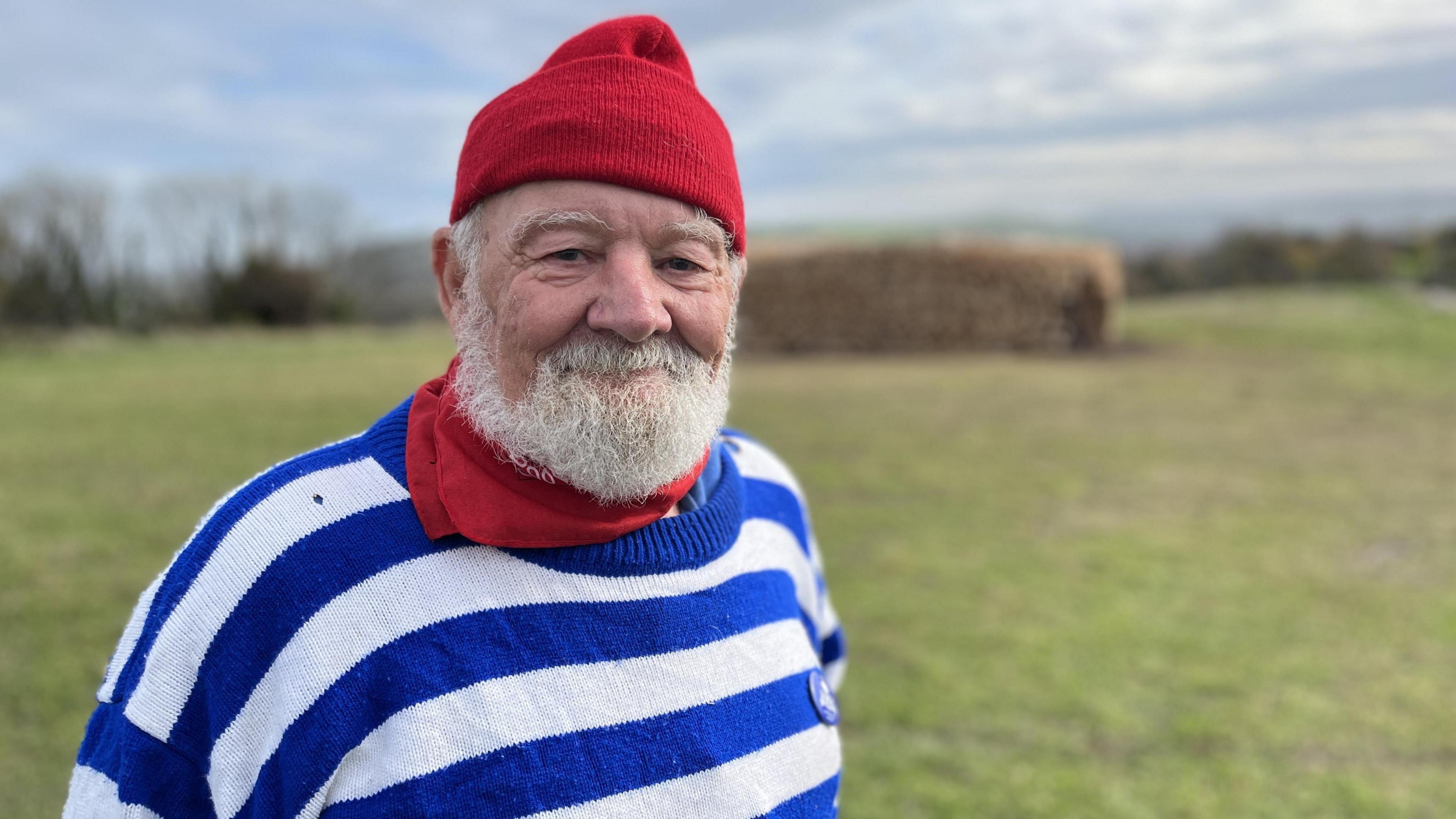 An elderly man with a white beard wearing a red woolly hat and neck scarf and a blue and white horizontal striped jumper is standing in a field