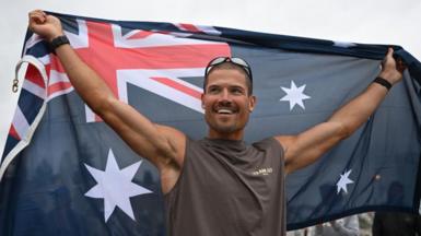 British ultra endurance runner William Goodge celebrates, holding an Australian flag, after crossing the finish line at Bondi Beach