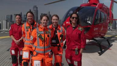 Group photo of the five female crew members on top of a building with the red helicopter behind them and a London skyline in the background