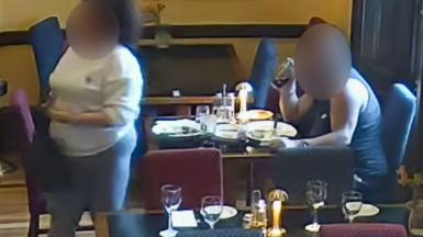 A man in a vest sat at a table with empty plates in front of him. He is drinking from a pint glass of coke. His face is blurred. On the other side of the table, a woman in grey leggings and a white sweatshirt is stood. Her face is also blurred. There are other restaurant tbales set with plates, cutlery and glasses around them.