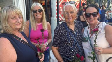 Four woman lining the street of Mumbles, Swansea. They are each holding a flower. They are smiling and looking at the camera.