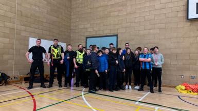 Cuid de na Poilis agus cuid de sgoilearan Sgoil MhicNeacail nan seasamh còmhla san talla spòrs agus iad air geama a chluich. 