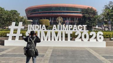 A man wearing a black pack takes a photo of a sign for the event, which says "Hashtag India AI Impact Summit 2026" in large white standalone letters. The sign sits in front of a round copper coloured building.