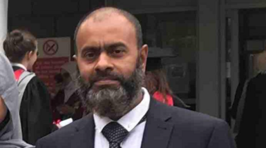 Man wearing suit, has short hair, balding, with grey/black beard. Wears a black suit with tie and white shirt