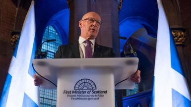 John Swinney, a bald man with glasses, dark suit and purple tie, gives a speech at a lectern saying "First Minister of Scotland". He is in the National Gallery of Scotland, a dark Gothic building, which has been lit in blue lights and two large Saltire flags flank him.