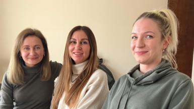 A picture of three women sitting down. On the left is a woman with light brown, shoulder-length hair, who is wearing a grey top and blue jeans. In the middle is a woman with longer brownish hair with blonde tints, wearing a white roll-neck jumper, and black trousers. On the right is a woman with blonde hair that is tied back, wearing a green hoodie and black jeans.