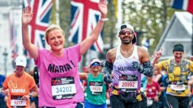 Maria Mulgrew running a competitive long-distance race. She is wearing a bright pink T-shirt decorated with her name "Maria!" in black letters, and black running shorts with white seams. She is smiling and has both arms raised above her head.