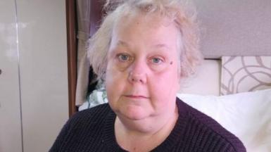 Donna is sitting in a motorhome-style space. She has light, slightly unkempt hair and wears a dark knitted jumper. In the background, there are white cushions, wood‑panelled cupboards and cream-coloured storage units.
