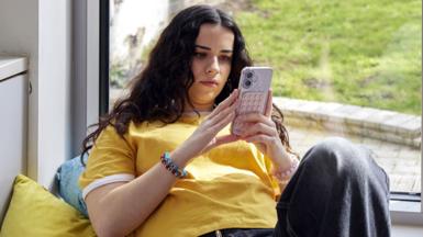 A young woman in yellow t-shirt reclines on cushions at home whilst scrolling her mobile phone