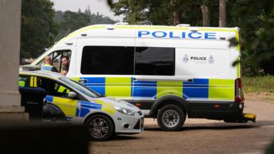 A large police van and a police car parked next to each other. Two officers - including one in the passenger seat and one standing outside - talking