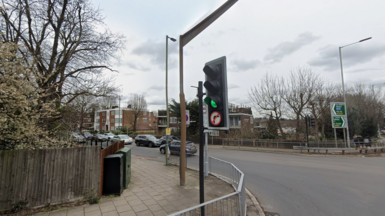 A general view of a pedestrian traffic light crossing on Holders Hill Road, with cars on the road and residential buildings and trees in the background.