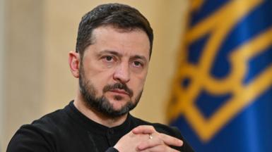 Zelensky looking off camera, with his hands clasped, and the Ukranian flag in the background