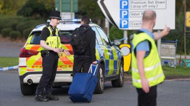 A man wheeling a suitcase talks to a police officer who is stood at the back of a police car, while another man in a hi-vis tabard is seen with is arm outstretched and appears to be giving directions.