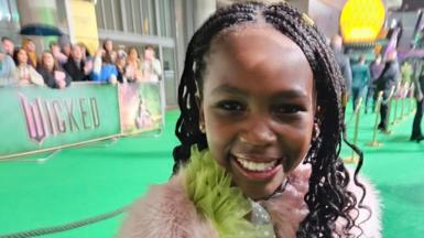 A young girl is stood on a green carpet whilst smiling at the camera. She is wearing a green dress and a pink fluffy coat.