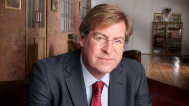 Edward Stourton pictured wearing a suit with a red tie in 2005, while posing for an indoor portrait with his hands clasped.