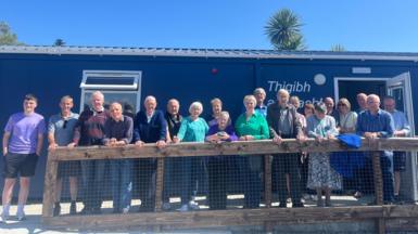 Dealbh de chuid de na daoine a bhios a' gabhail pàirt sa chluba gach Diciadain agus iad nan seasamh taobh a-muigh an togalaIch a bhios iad a' cleachdadh airson coinneachadh. Tha là math ann 's an adhar gorm. Tha gach neach a' coimhead uabhasach toilichte. 