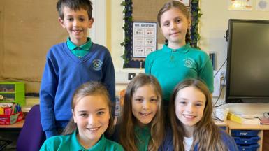 Five pupils wearing green polo shirts in two rows in a classroom with two pupils standing at the back and three girls sitting in front of them. There is one boy standing, wearing a blue jumper. All the children are smiling. 
