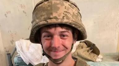 James Witton smiles for the camera, he is wearing a military helmet with camouflage covering. Behind him is a bed with  a bare mattress and some objects on it   