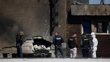 Four men in police vests, as well as a man in a white hazmat suit, a helmet and a face mask, are seen stood next to a building which has a hole in its side following the drone hit