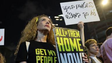 A woman stands at a protest holding a placard reading "All of them home now"