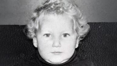 A black and white image of a young boy with blond hair, looking into the camera with a smile