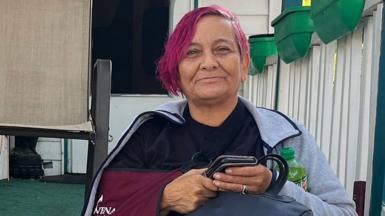 Head and shoulders shot of Rachel Oviedo, 57, sitting on her porch with pink hair, wearing a grey top and holding her phone