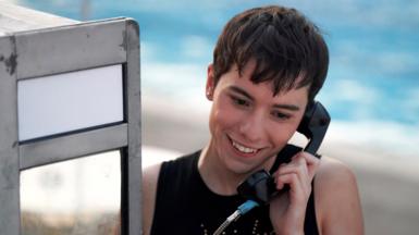 Ellis Howard as Byron in What It Feels Like For A Girl, using a pay phone and smiling.
