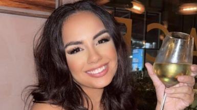 A young woman with long, dark hair and brown eyes smiles at the camera with a glass of white wine in her hand. She appears to be sitting in a booth at a bar or restaurant