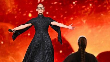 A female singer at Eurovision stands in a black dress with her arms out in front of a fiery backdrop.