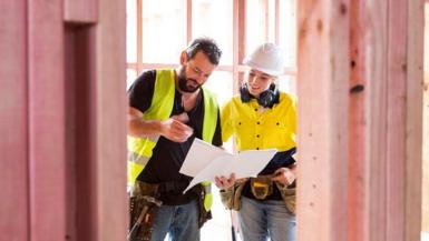 Bearded man and woman in high vis jackets look at plans for new home in a building site