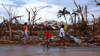 Three people walking past broken trees and blown down houses left by the passage of hurricane Melissa.
