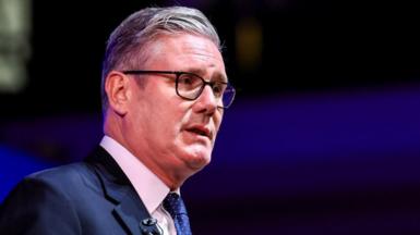 UK Prime Minister Keir Starmer sideways on in a navy blue jacket with a white shirt, navy tie with white polka dots and a small black mic on his lapel. He is also wearing black glasses.
