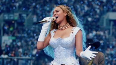 Beyoncé close-up in an all-white outfit gesturing while holding the microphone as she sings on stage