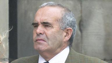 A man with short grey hair and black eyebrows pictured outside. He is wearing a green checked jacket and a shirt and tie