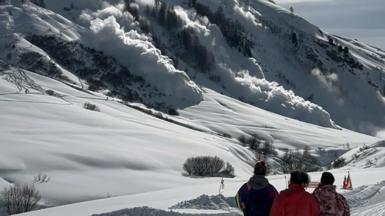 Three people watch in foreground as avalanche barrels down mountainside ahead, from left to right