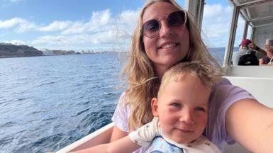 Megan has long blonde wavy hair and is taking a selfie of herself and Edward, who is sitting on her lap. They are on a boat in the sunshine, with white hotels in the background along the coastline.