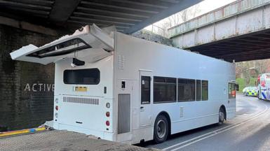 White double decker bus with roof ripped off after it hit a low bridge. A police and fire engine is seen past the bridge on a bend further up the street.