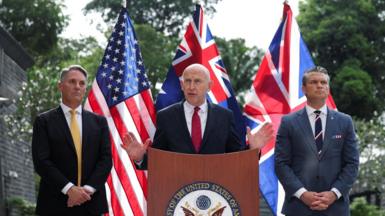 John Healey speaks and gestures with his hands at a podium at the US embassy, flanked by Richard Marles and Pete Hegseth. The US, UK and Australia flags are posted behind them.