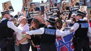 Protesters holding up placards with Keir Starmer's face printed on them being held back by police in Golders Green today 3oth April 2026