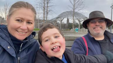 A selfie-style family photo of a woman and a man either side of a young boy, who is sticking his tongue out while smiling. They are all looking into the camera smiling.