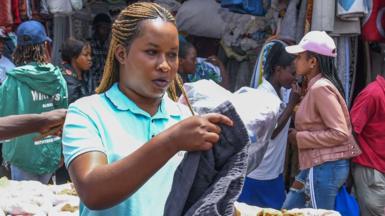 A clothes vendor with long braids and in a blue T-shirt (L) holds up black trousers at Gikomba market in Nairobi, Kenya. Behind her can be seen people walking past other stalls -September 2023.